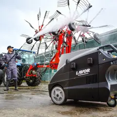 Un hombre con mono y gorra usa una hidrolimpiadora para limpiar un rastrillo agrícola. Así funciona una hidrolimpiadora.