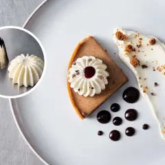 Ein Stück Kuchen mit aufgespritzteter schwerer Sahne, Schokoladensauce und Kakaonibs. Daneben ein Spritzbeutel mit einer Sahnetülle und ein Beispiel für aufgespritztte Sahne.