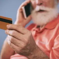 Starszy mężczyzna z brodą trzyma kartę i rozmawia przez telefon, zastanawiając się, dlaczego bank zablokował kartę.