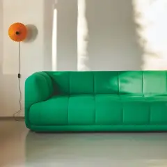 Ein leuchtend grünes Sofa steht vor einer weißen Wand, an der eine orangefarbene Lampe hängt. Sonnenlicht wirft Schatten. Mette hay.
