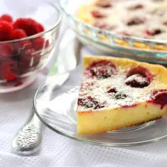 Une part de clafoutis framboise, saupoudrée de sucre glace, à côté d'un bol de framboises fraîches.