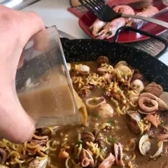 Vertiendo caldo en una paellera con fideos, calamares y almejas. Así se hace fideuá.