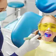 Dziecko w okularach ochronnych uśmiecha się, gdy dentystka utwardza lakowanie zębów. Czy lakowanie zębów jest szkodliwe? Nie, to bezpieczna profilaktyka.