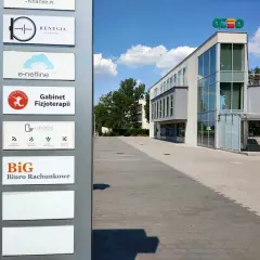 Tablica z nazwami firm, w tym "BiG Biuro Rachunkowe", przed nowoczesnym budynkiem biurowym.