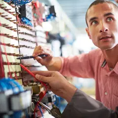 Dwóch mężczyzn pracuje przy panelu elektrycznym, jeden trzyma miernik.