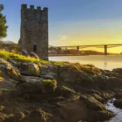 Torre medieval junto a un río al atardecer, con un puente al fondo. Imagina el tren Madrid Ferrol pasando por allí.