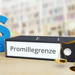 Ein Ordner mit der Aufschrift "Promillegrenze" steht neben Paragraphen-Symbolen und einer Waage. Dies symbolisiert die rechtlichen Aspekte der Promillegrenze im Straßenverkehr.
