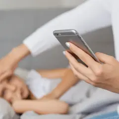 Kobieta z telefonem w dłoni sprawdza gorączkę dziecku leżącemu w łóżku.