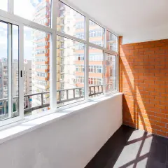 Szklana loggia z widokiem na blokowisko. Jasny balkon z białymi oknami i ceglaną ścianą.