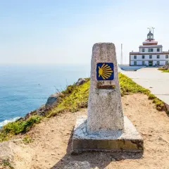 Kamienna wskazówka z symbolem muszli i napisem "0.000 KM" na trasie Camino Portugues. W tle biała latarnia morska i błękitne morze.