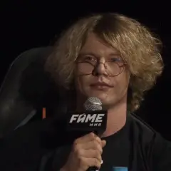 Natan Marcon z burzą blond włosów i okularami, trzymający mikrofon FAME MMA. Jego wiek jest trudny do określenia, ale wygląda na młodego mężczyznę.