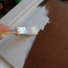 Main d'une personne appliquant une couche de peinture claire sur un meuble en bois verni.