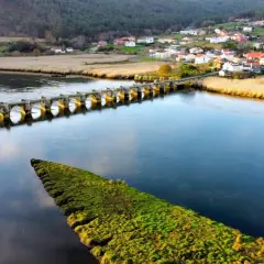Un puente de piedra con arcos cruza un río azul, conectando un pueblo pintoresco con campos verdes. El puente, conocido como Ponte nafonso, se alza majestuoso sobre el agua.