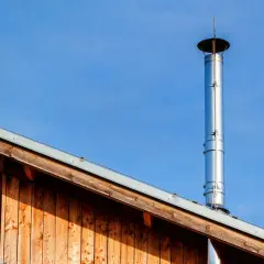 Błyszczący, metalowy komin zewnętrzny do kozy, zamontowany na drewnianym dachu, na tle błękitnego nieba.