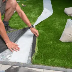 Mann verlegt Kunstrasen, legt die Nahtstelle frei und bereitet sie für das Verkleben vor. Sandfüllung liegt bereit.