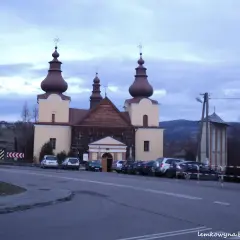 Cerkiew łemkowska z charakterystycznymi kopułami, otoczona samochodami, na tle pagórkowatego krajobrazu.