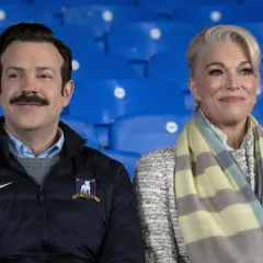 Die Besetzung von Ted Lasso, Jason Sudeikis und Hannah Waddingham, sitzt lächelnd auf blauen Tribünen.