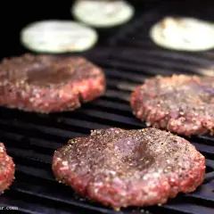 Vier rohe Burger-Patties, die darauf warten, dass man sie selber macht, brutzeln auf einem Grill. Zwiebelringe liegen im Hintergrund.