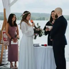 Panna młoda z mikrofonem i bukietem mówi podczas ceremonii. Obok stoi pan młody, urzędniczka i świadkowa. To ślub jednostronny nad jeziorem.
