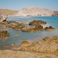 Rocas volcánicas emergen de las aguas tranquilas de la cala de la media luna, con montañas áridas al fondo bajo un cielo azul.