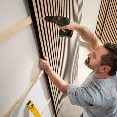 Man skruvar fast en akustikpanel på väggen med en borrmaskin. En vattenpass ligger redo.