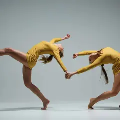 Deux danseuses en tenue jaune, mains jointes, en mouvement synchronisé sur fond gris.