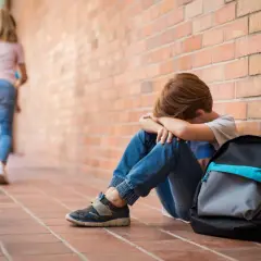 Chłopiec siedzi przy ceglanej ścianie, przytulając kolana. Inne dzieci biegną obok, nie zwracając na niego uwagi. To obraz samotności i fobia społeczna u nastolatków.