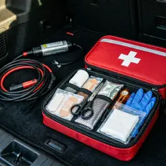 Czy apteczka jest obowiązkowa w samochodzie? W bagażniku leży czerwona apteczka z białym krzyżem, obok kable rozruchowe.