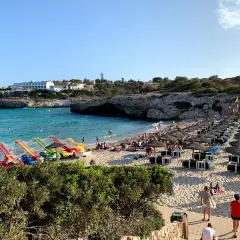 Słoneczna plaża Cala Domingos tętni życiem. Woda kusi turkusowym blaskiem, a piasek ugina się pod ciężarem leżaków i parasoli. Dzieci bawią się na kolorowych zjeżdżalniach.