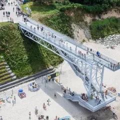 Nowa kładka na plaży Trzęsacz ułatwia dostęp do morza. Ludzie spacerują po niej, podziwiając widoki.