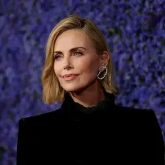 Charlize Theron, bekannt für ihre Rollen in Filmen wie "Mad Max: Fury Road", posiert vor einem lila Hintergrund.