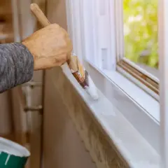 Malowanie parapetu okna plastikowego białą farbą. Puszka z farbą do malowania okien plastikowych stoi obok.