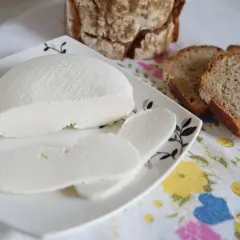 Świeży ser typu bufala, pokrojony w plastry, obok kromek chleba na kwiecistym obrusie.
