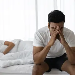 Uomo seduto sul letto con le mani sul viso, preoccupato per l'eiaculazione precoce. La donna dorme nel letto alle sue spalle.