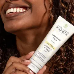 Une femme souriante tient un tube de gommage visage éclat. Ce produit sert à lisser et illuminer la peau, révélant sa beauté naturelle.