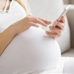 Kobieta w 24. tygodniu ciąży przegląda coś na telefonie, siedząc na kanapie.