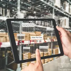 Ręce trzymają tablet z widokiem magazynu, pokazujący alerty "Running Out Of Stock" i czas kompletacji zamówienia. To przykład nowoczesnego zarządzania łańcuchem dostaw.
