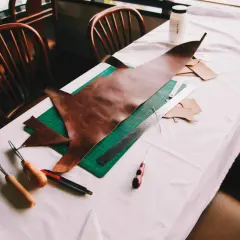 Sur une table recouverte d'une toile blanche, des outils de maroquinerie et des pièces de cuir sont disposés. On y voit un grand morceau de cuir brun, une planche à découper verte, une règle, un cutter, et des outils pour travailler le cuir, suggérant ...