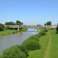 Metalowy most kratownicowy nad rzeką, otoczony zielonymi brzegami i drzewami.