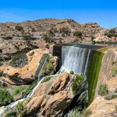 Cascada en el pantano de Elche, con rocas cubiertas de musgo y vegetación verde. Personas observan el agua.