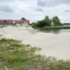 Piasek na plaży w Kamieniu Pomorskim czeka na budowę. W tle zielone budynki i spokojna woda.