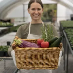 Une opératrice certiphyto souriante tient un panier rempli de légumes frais : aubergine, tomates, concombres et romarin, dans une serre.
