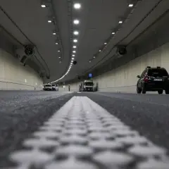Wnętrze tunelu z widocznym ruchem pojazdów i pracami drogowymi. Widać białą linię pasów ruchu i czarny samochód. Odstęp w tunelu jest wyraźnie zaznaczony.