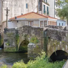 Puente de piedra con arcos sobre río, cruz de piedra y edificios históricos. Un lugar encantador que ver en Caldas de Reis.