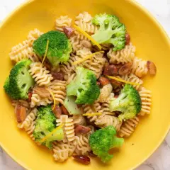 Macarrones con brócoli, trozos de salchicha y ralladura de limón en un plato amarillo.