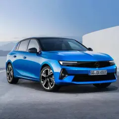 Błękitny opel elektryczny z czarnym dachem, zaparkowany na betonowej nawierzchni, z górzystym krajobrazem w tle.