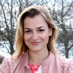 Uśmiechnięta Ines Lutz, znana z roli w "Der Bergdoktor", w różowej kurtce na tle drzew.