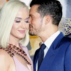 Orlando Bloom szepcze coś do ucha Katy Perry, która ma na sobie różową sukienkę i gruby, złoty naszyjnik.