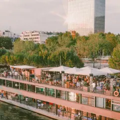 Un bateau-mouche animé sur la Seine, avec des gens faisant la fête sur le pont. Le soleil brille sur un gratte-ciel, créant un effet de "mazette" lumineux.