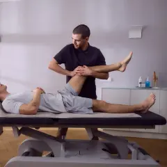 Physiotherapeut dehnt das Bein eines Patienten auf einer Liege. Die Knie-Schiene nach OP wird sanft bewegt, um die Beweglichkeit wiederherzustellen.